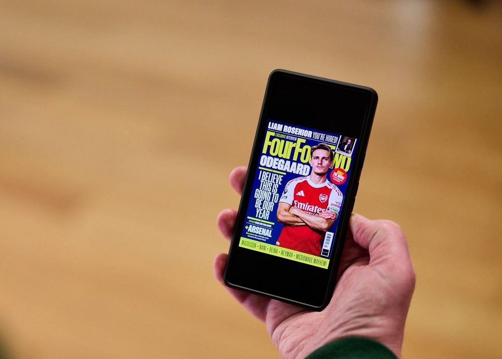 A close-up of a person's hand holding a smartphone. The screen displays a digital issue of "FourFourTwo" magazine featuring football player Martin Ødegaard on the cover.