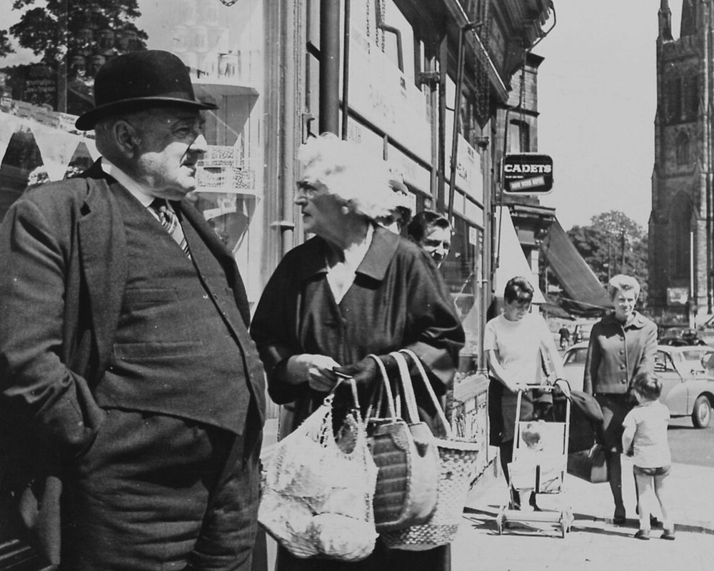 A black & white photo of shoppers of shoppers on a street in Newcastle in the Sixties. We can see a man in a suit and hat, a lady with shopping bags and two ladies with children.