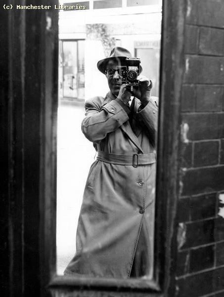 A black and white shot of photographer Lawrence Kaye standing in front of a full-length mirror, photographing himself. He is wearing a trenchcoat and hat.