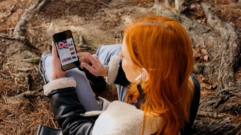 A young woman is sitting beneath a tree reading a digital magazine on a smartphone.