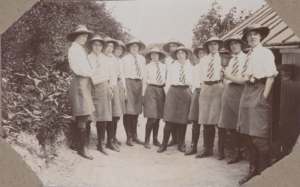 Sepia photo of several young women in uniforms (shirts, ties, skirts, hats, boots) standing outdoors in a line with bicycles.