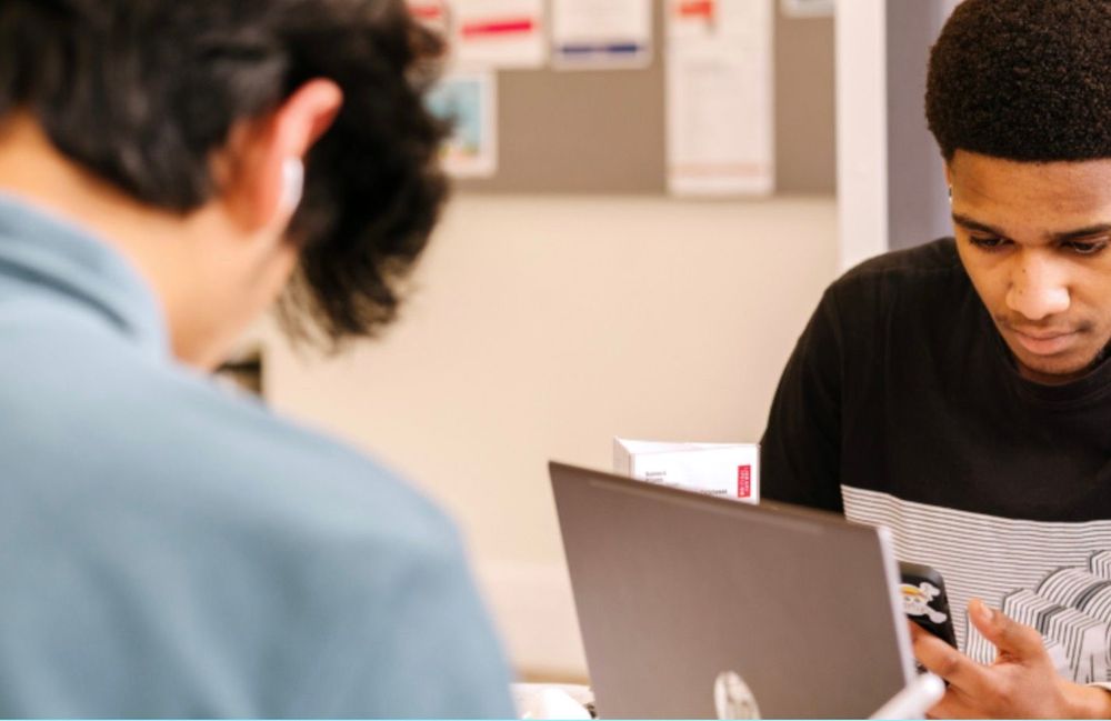 Two young men around a table at the library. One has his laptop and is looking at his phone, the other has a tablet stylus in his hand. They are both concentrating on their tasks