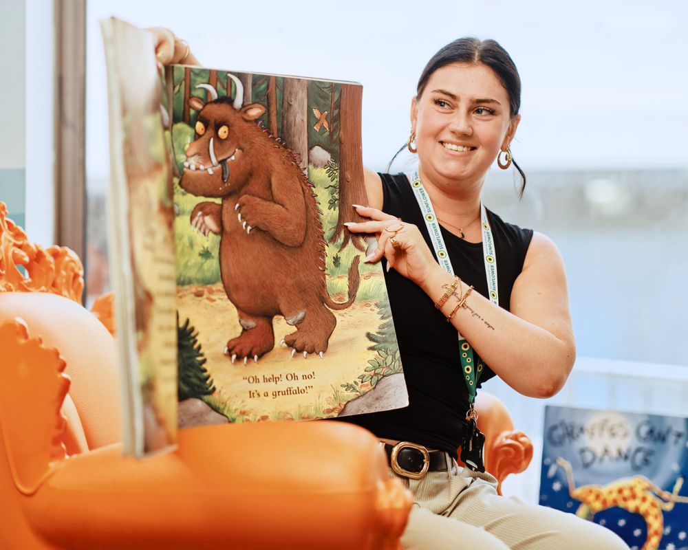 A female library staff member smiles as she reads a story to a group of children.