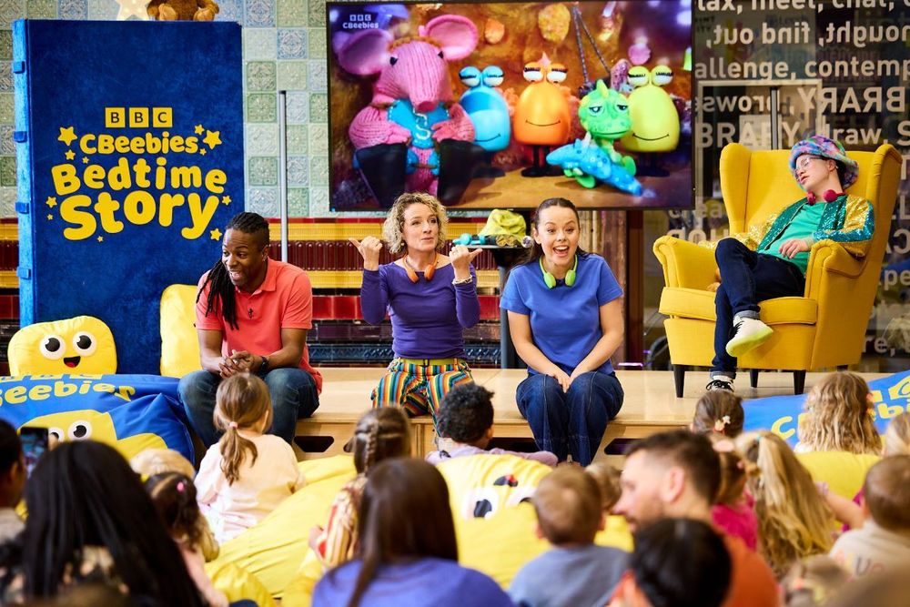CBeebies presenters speak to a crowd of children and parents at a colourful event in Leeds Central Library. In the background a stand has the words: 'BBC Children's Bedtime Story'.