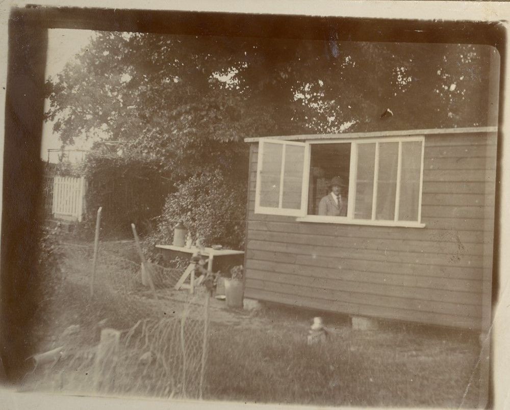 Sepia photo of a wooden shed with an open window; a person looks out. There are garden tools in foreground.