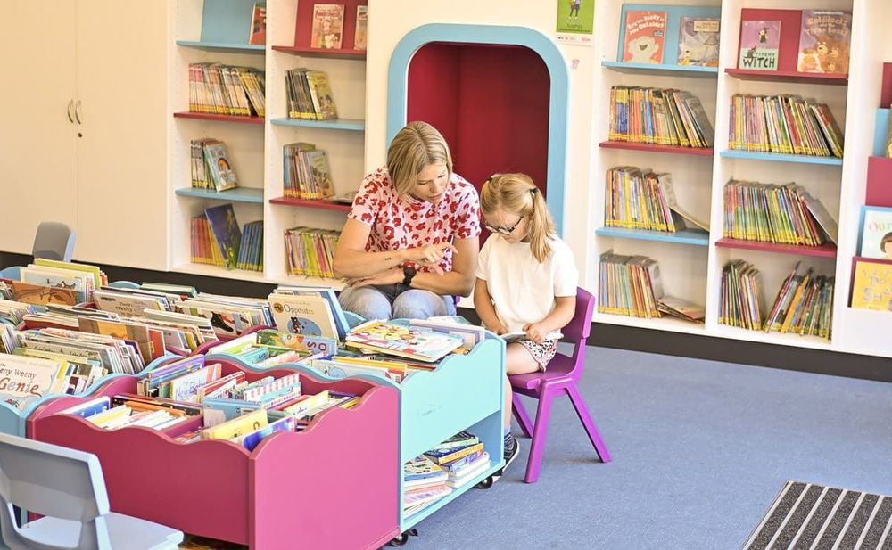 An adult kneels beside a young child in a colorful children's library section. They are looking at a book together near low bookshelves and a bright red reading nook.