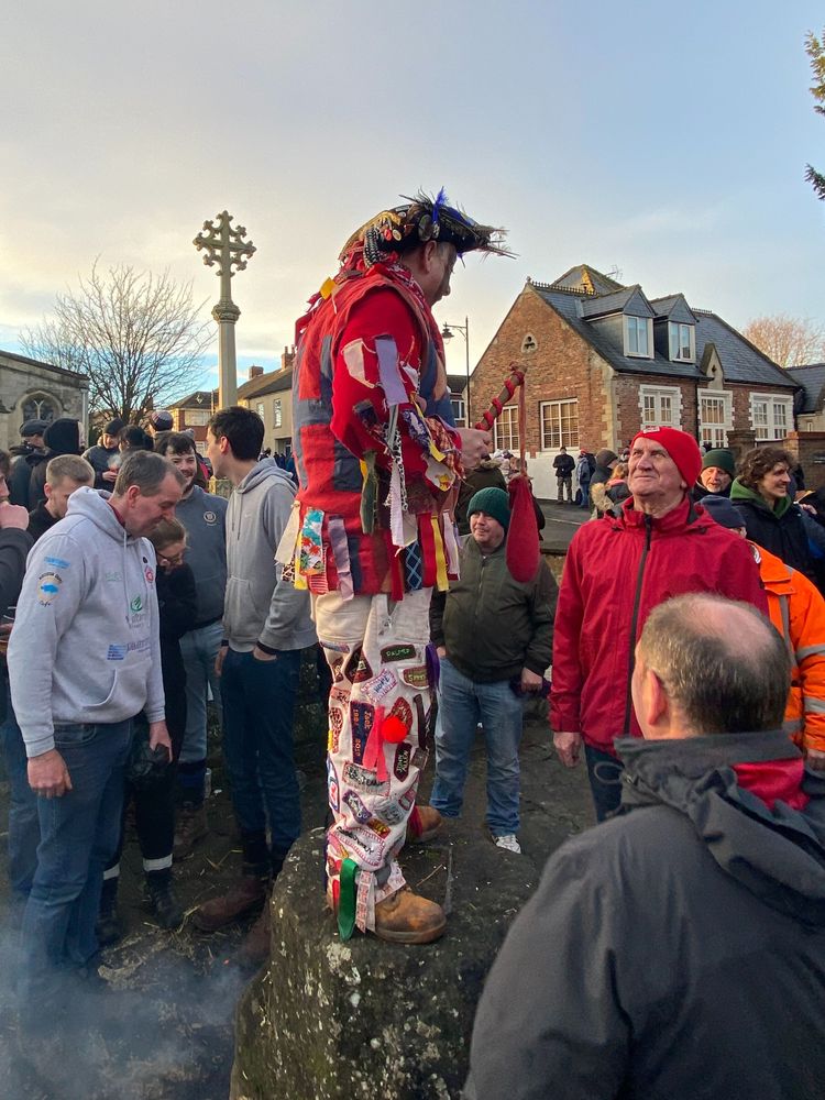 A man in harlequin jacket, white trousers and tricorn hat surrounded with smoke, standing on a stone.