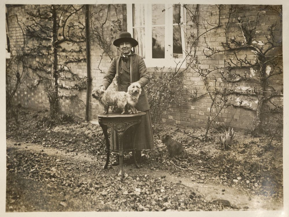 Sepia photo of a woman in a hat, in a garden with three small dogs and a house.
