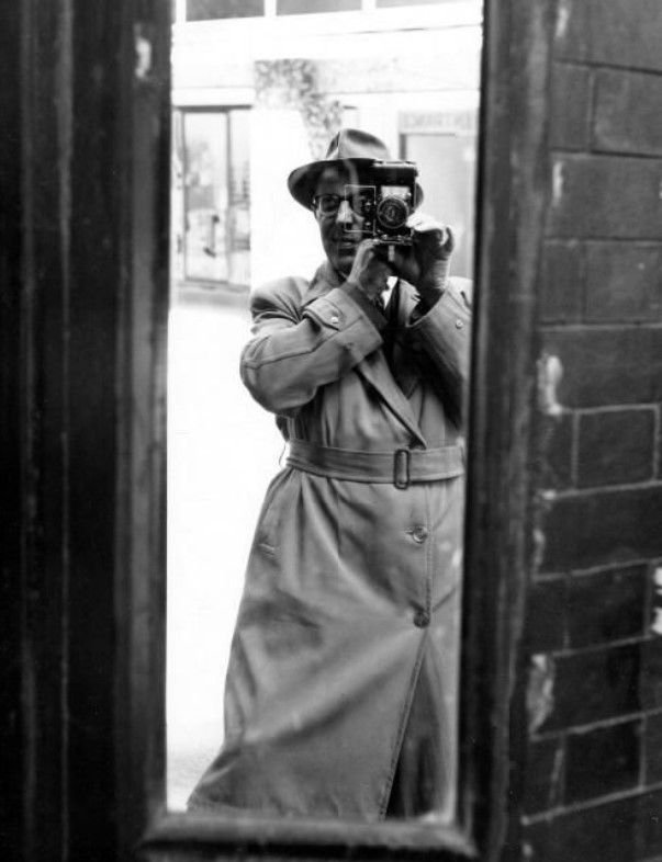 A black and white shot of photographer Lawrence Kaye standing in front of a full-length mirror, photographing himself. He is wearing a trenchcoat and hat.
