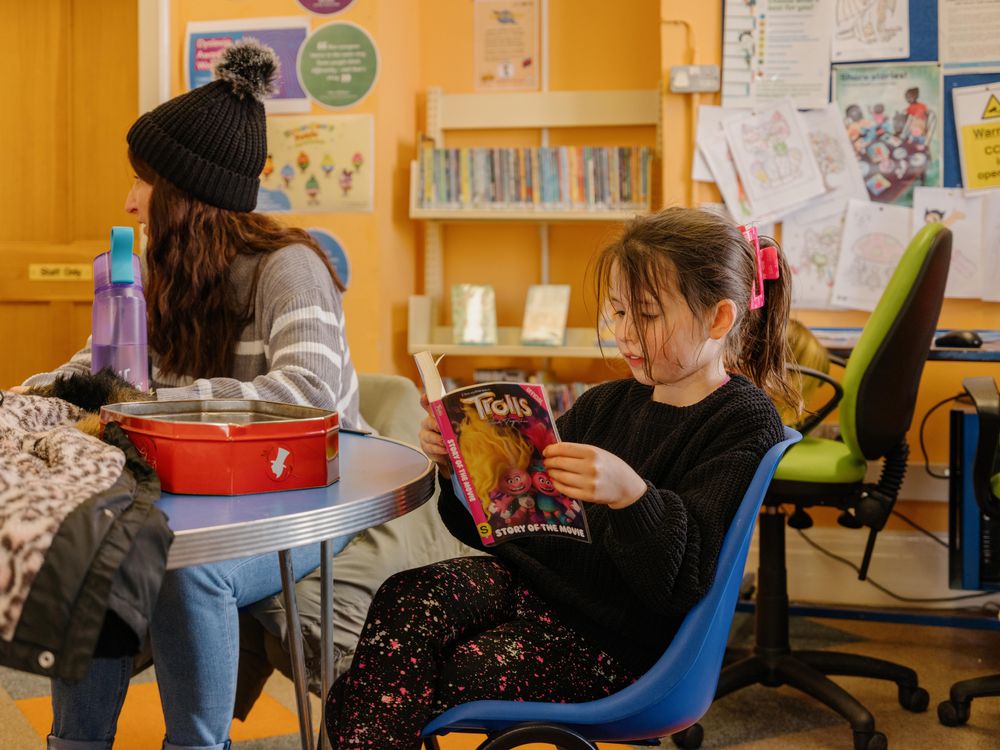 A young girl sits at a blue table in a library, intently reading a "Trolls" movie storybook.