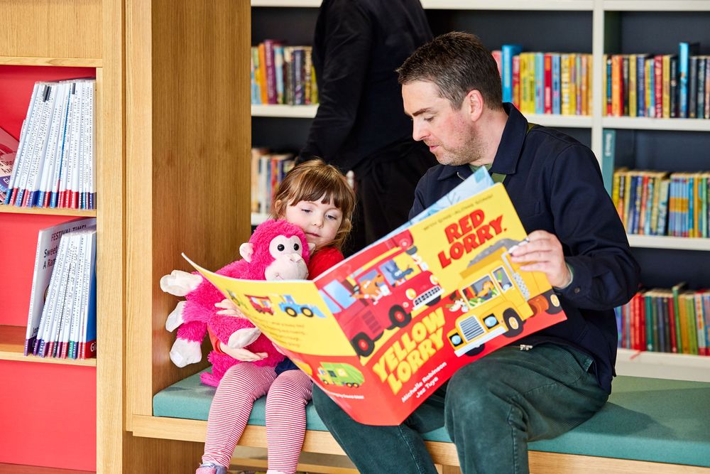 An adult and a young child sit together on a library bench, reading a large picture book titled Yellow Lorry, Red Lorry. The child is holding a pink plush monkey.