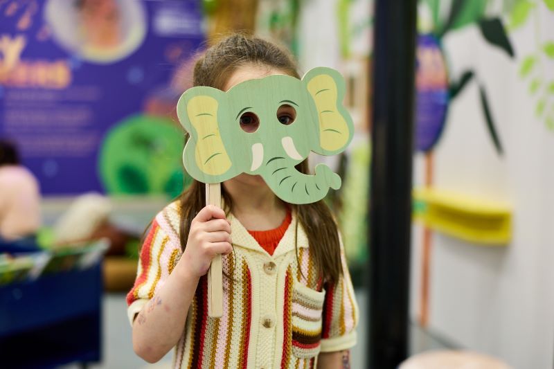 A child in a library holds an illustrated elephant mask in front of their face.