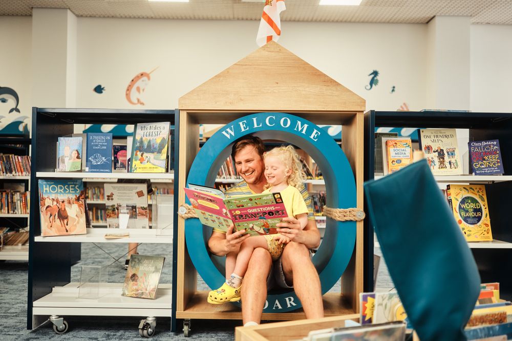 An adult and child laugh together while reading a book about transport inside a unique, circular blue reading nook in Jersey Library.