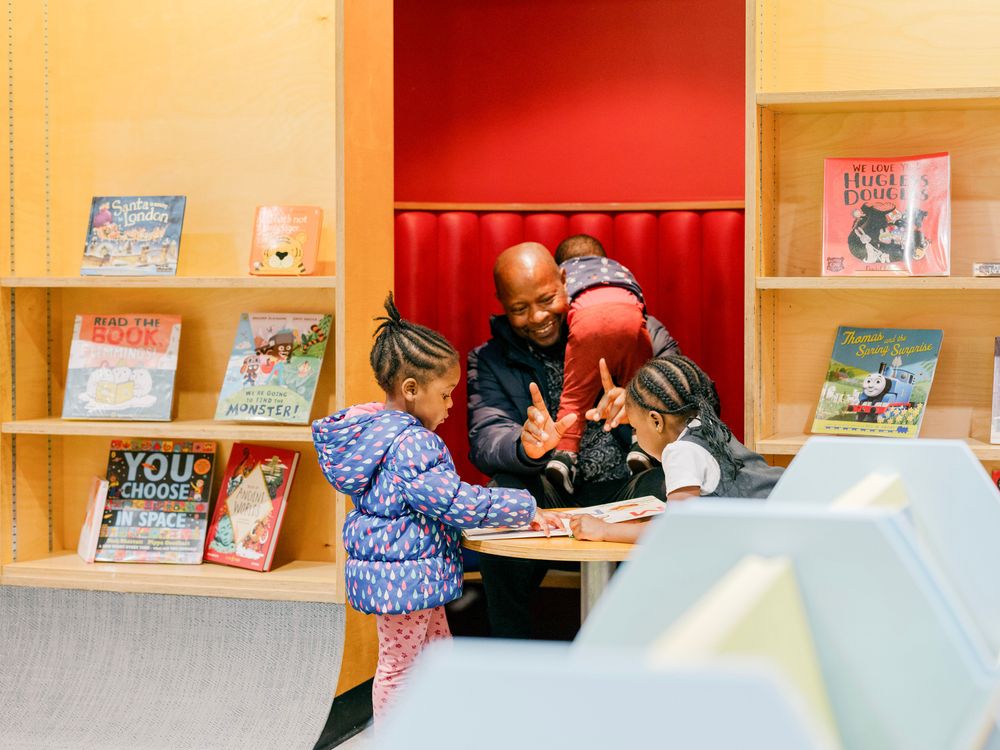 An adult and three children are having fun around a small, low table in a bright and welcoming children's library area.