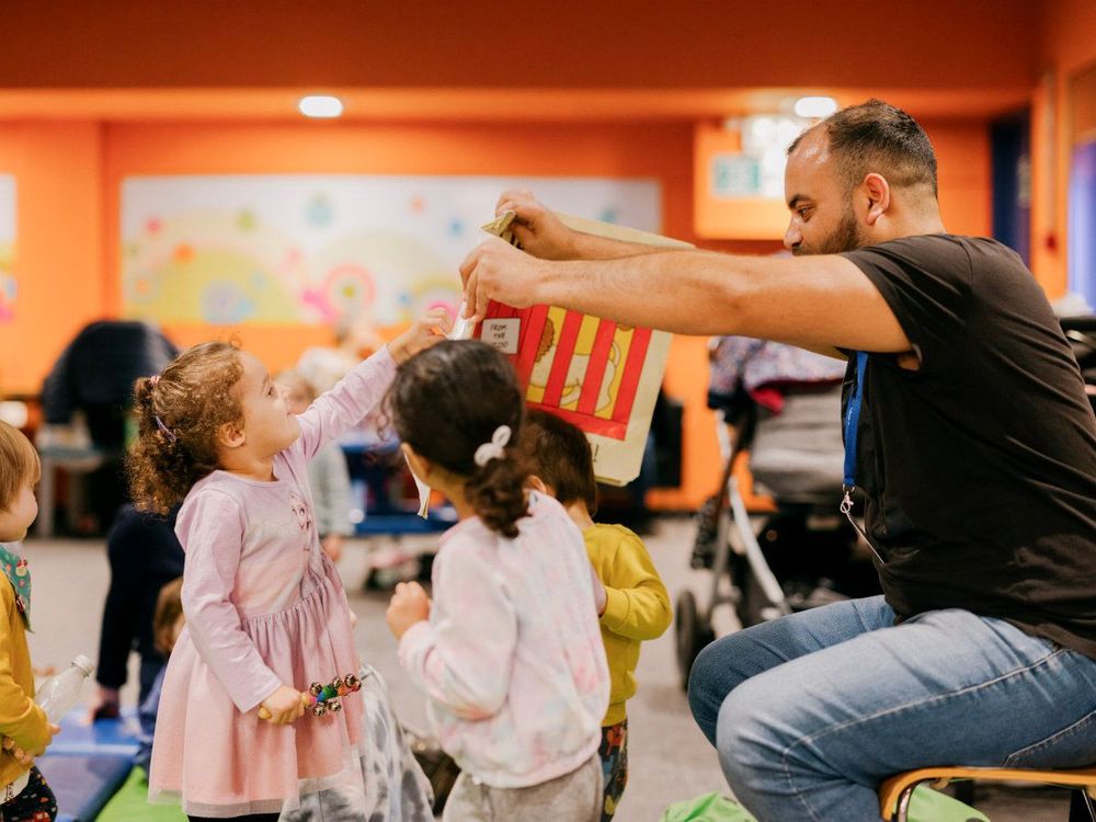 A librarian holds a book open infront of some children. One of the children is pointing at the book.