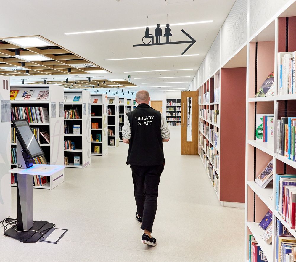 In a brightly-lit library a figure walks past shelves full of books. They are wearing a top which has 'Library Staff' printed on the back.