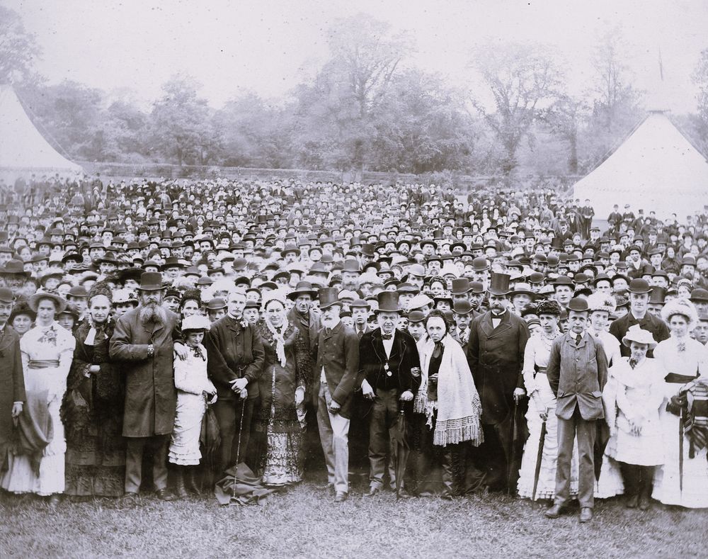 A sepia photo of a large crowd of several hundred people posing for the camera. The people are dressed formally, with men in suits and women in dresses. All the men and women wear hats or bonnets.